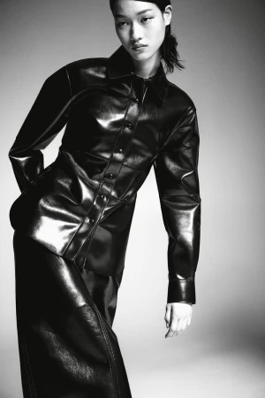 Monochrome portrait of a female model in a shiny black, long-sleeved shirt and matching skirt, set against a plain grey background.