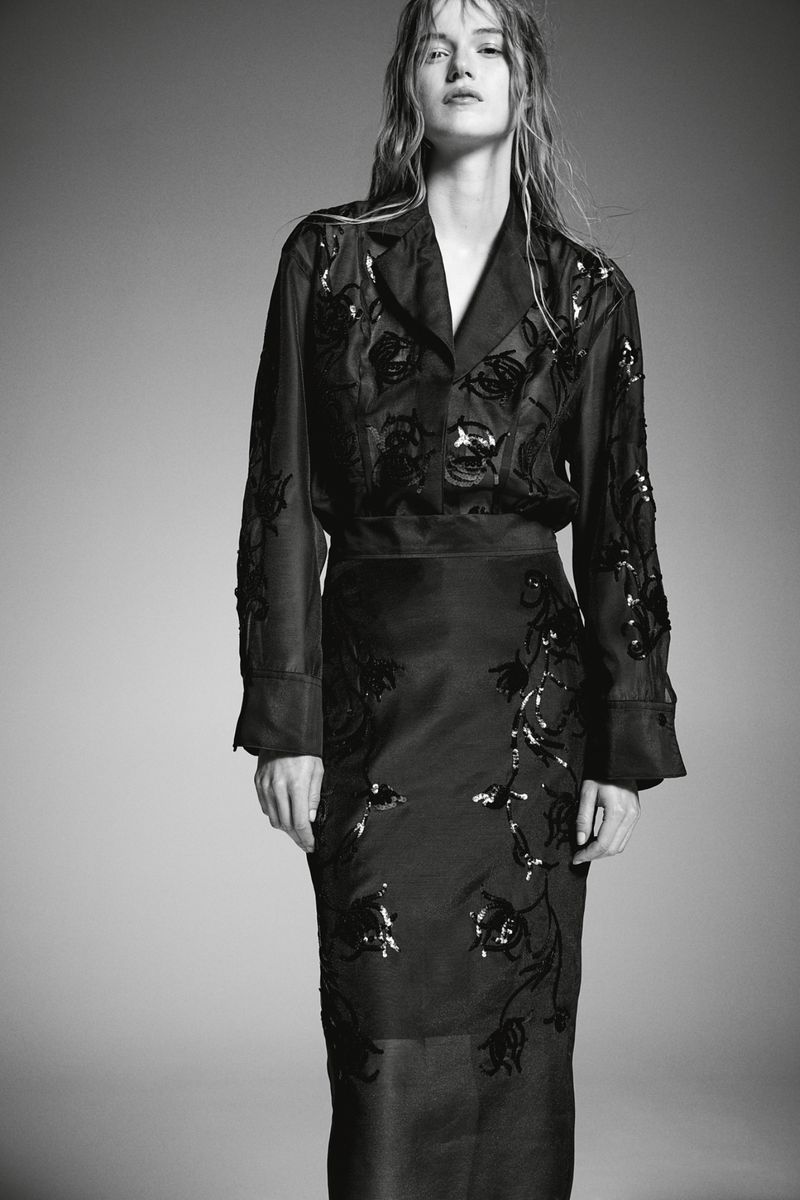 Woman wearing black floral embellished jacket and skirt posing in front of grey backdrop in studio lighting for fashion shot.