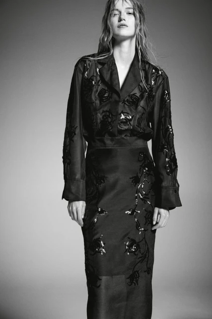Woman wearing black floral embellished jacket and skirt posing in front of grey backdrop in studio lighting for fashion shot.