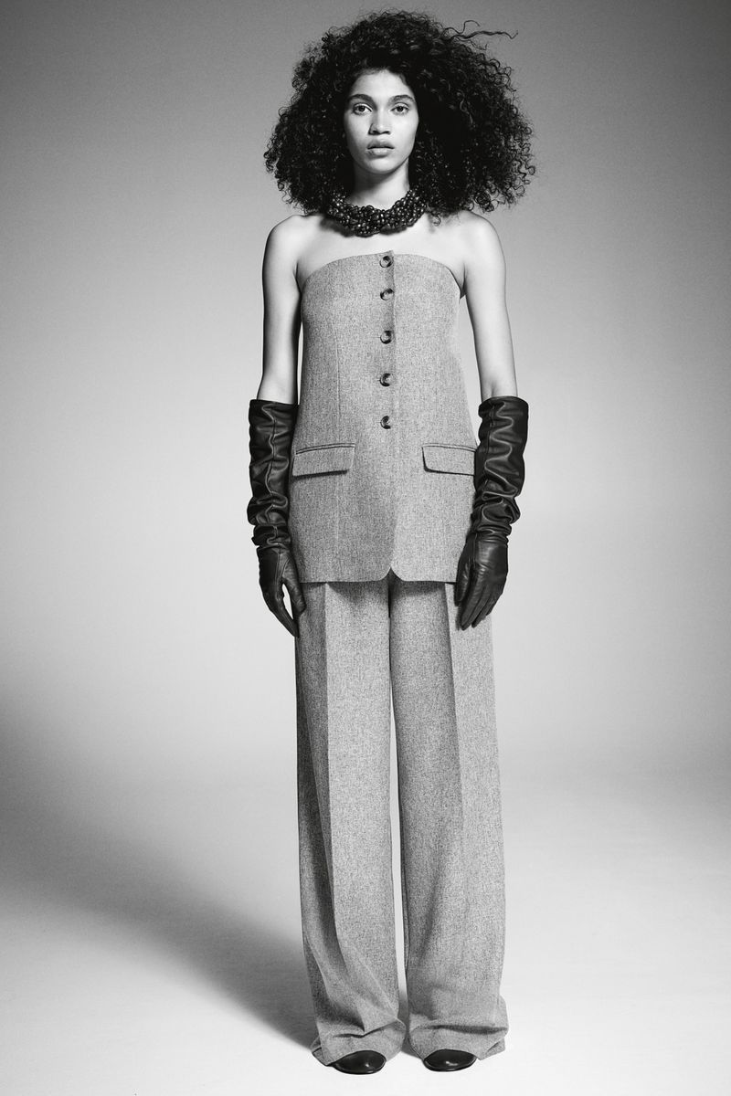 A slender woman with dark curly hair poses in a grey wool tube top and wide-leg pants, complemented by black leather gloves and necklace, indoors.