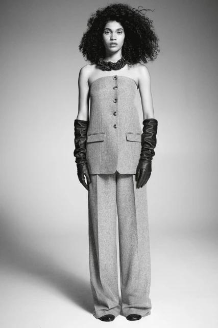 A slender woman with dark curly hair poses in a grey wool tube top and wide-leg pants, complemented by black leather gloves and necklace, indoors.