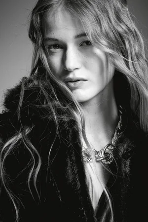 Close-up portrait of a woman with long wavy hair wearing a textured dark jacket and a chunky silver chain necklace against a grey backdrop.