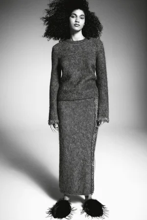 Model with voluminous curly hair wearing a textured grey knit sweater and skirt against a seamless white background.