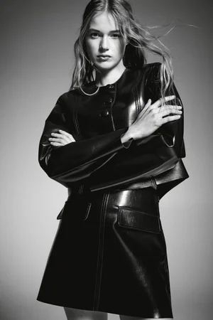 Model with long hair blowing in the wind wearing a black leather jacket and skirt against a grey background
