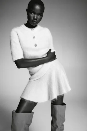 Model wearing a fuzzy white short-sleeve sweater, skirt, and boots against a gradient grey studio background.
