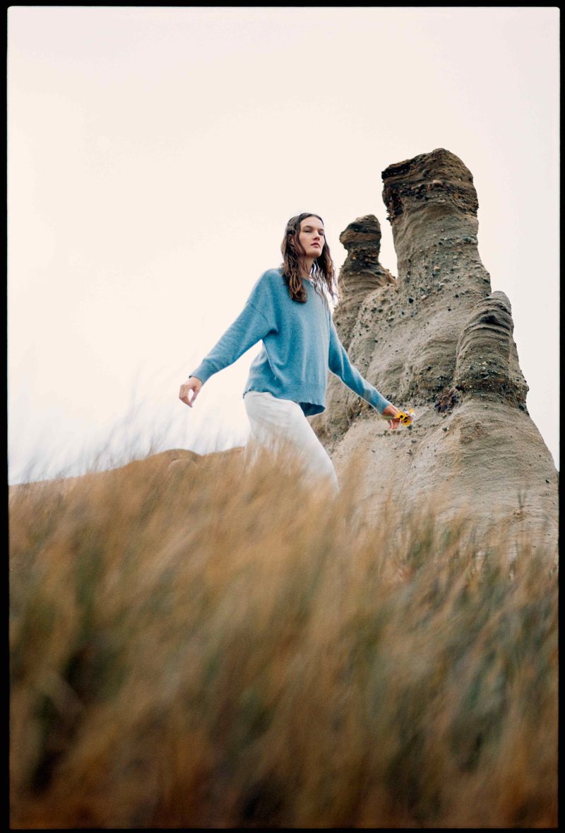 A young woman in blue long-sleeve shirt and white pants walks through tall golden grass near unique rock formations on a cloudy day.