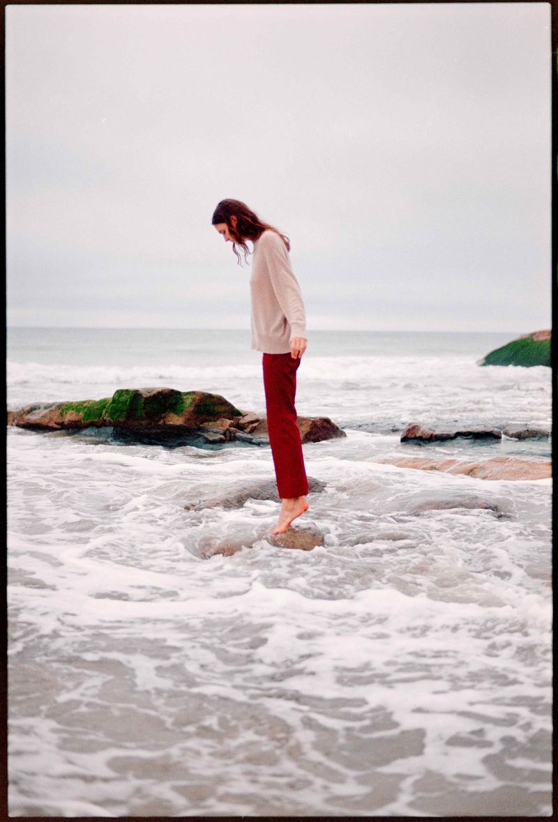 A woman in a sweater and maroon pants stands barefoot on rocks in the ocean, looking down at the water on an overcast day.