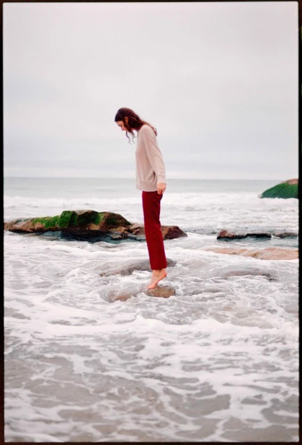 A woman in a sweater and maroon pants stands barefoot on rocks in the ocean, looking down at the water on an overcast day.
