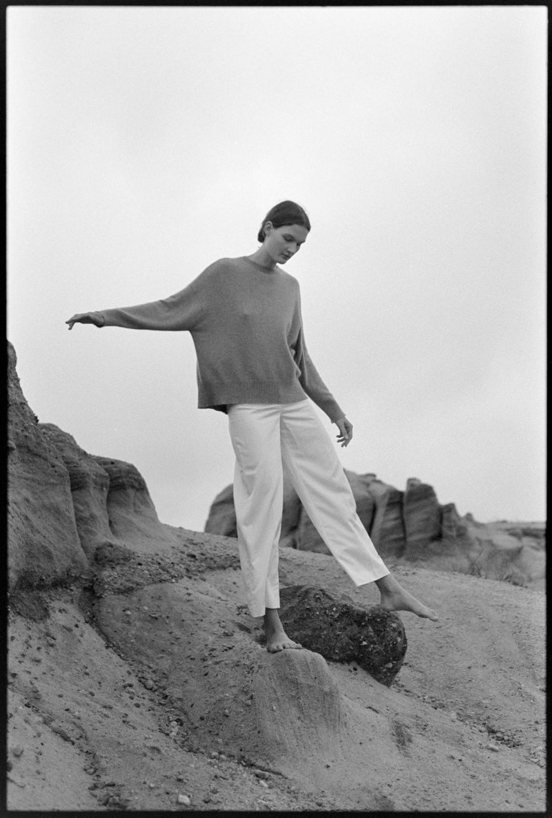 Woman in casual sweater and white pants balances barefoot on rocks in an outdoor setting under an overcast sky, creating a serene mood.