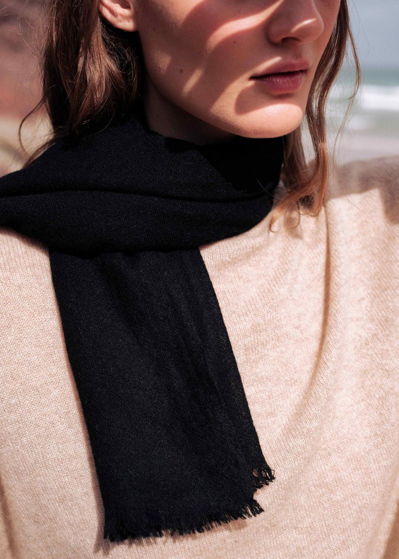 Close-up shot of an adult woman wearing a black scarf and tan sweater, standing outdoors with a blurred seascape in the background.