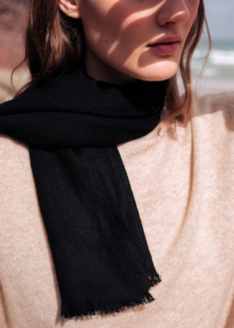 Close-up shot of an adult woman wearing a black scarf and tan sweater, standing outdoors with a blurred seascape in the background.