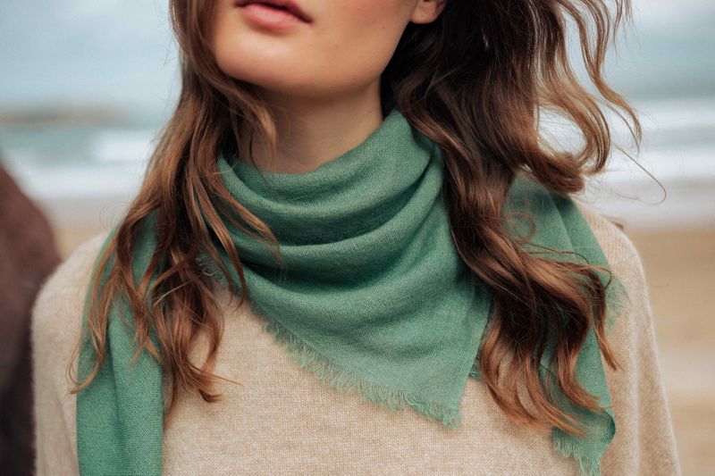 Close-up of a fashionable adult woman wearing a green fringed scarf and a tan sweater, standing on a beach with ocean in the background.