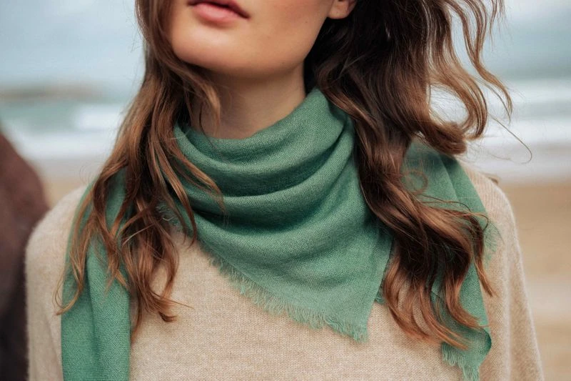 Close-up of a fashionable adult woman wearing a green fringed scarf and a tan sweater, standing on a beach with ocean in the background.