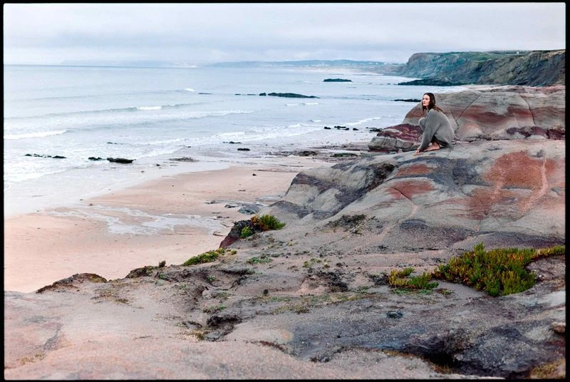Adult woman sits on rocky cliff overlooking ocean. Person in casual clothing enjoys coastal view. Calm sea meets rugged shoreline.