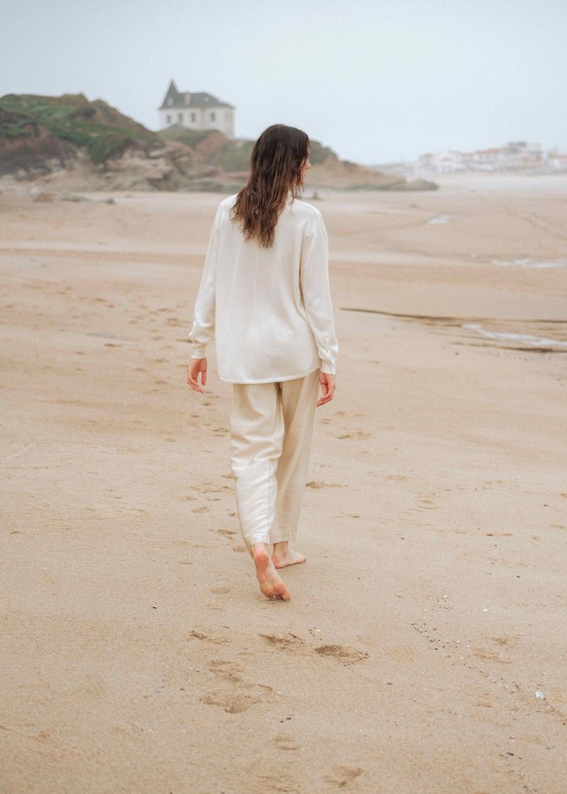 A woman in casual clothing walks barefoot on a sandy beach near a coastline with a building on the horizon, during an overcast day.