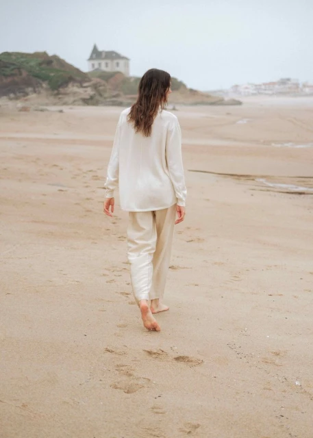 A woman in casual clothing walks barefoot on a sandy beach near a coastline with a building on the horizon, during an overcast day.
