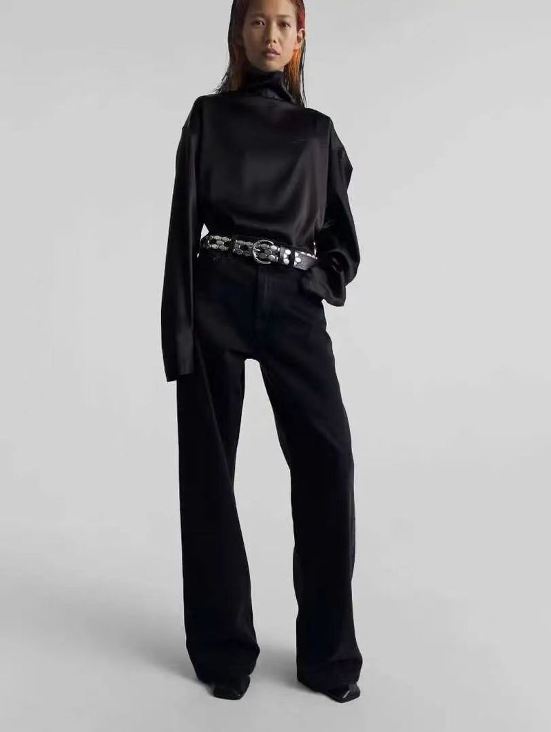 Studio shot of a young adult posing in a long-sleeve black top, dark pants, and a detailed silver belt against a neutral background.