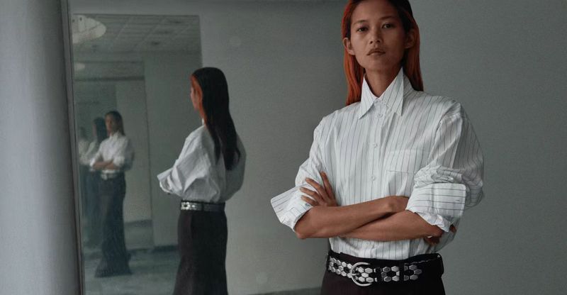 Adult woman with auburn hair wearing a button-down shirt with pinstripes and a studded belt, standing indoors with arms crossed.