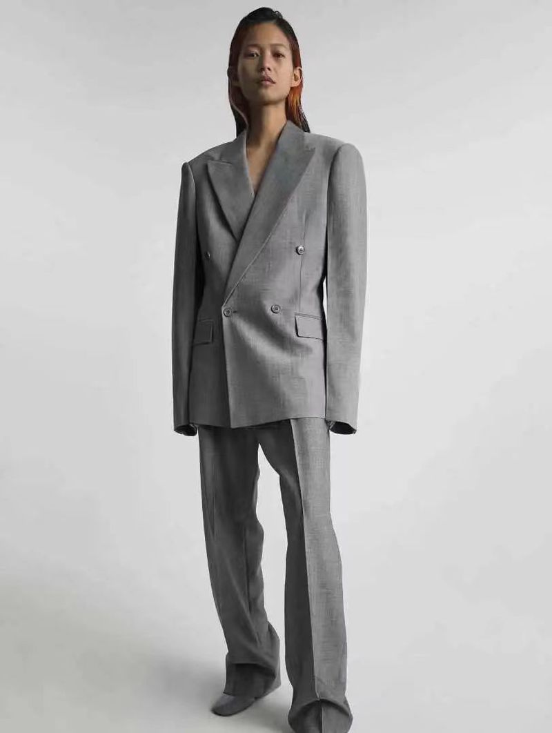 An Asian woman poses in a gray double-breasted suit with wide-leg trousers against a white background in a fashion studio setting.