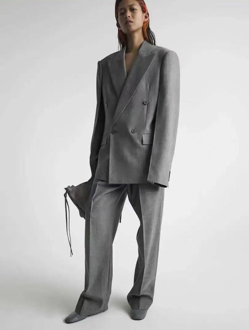 An adult stands in a studio wearing a grey wool suit with wide legs and oversized blazer. She holds a grey bag with drawstring. 