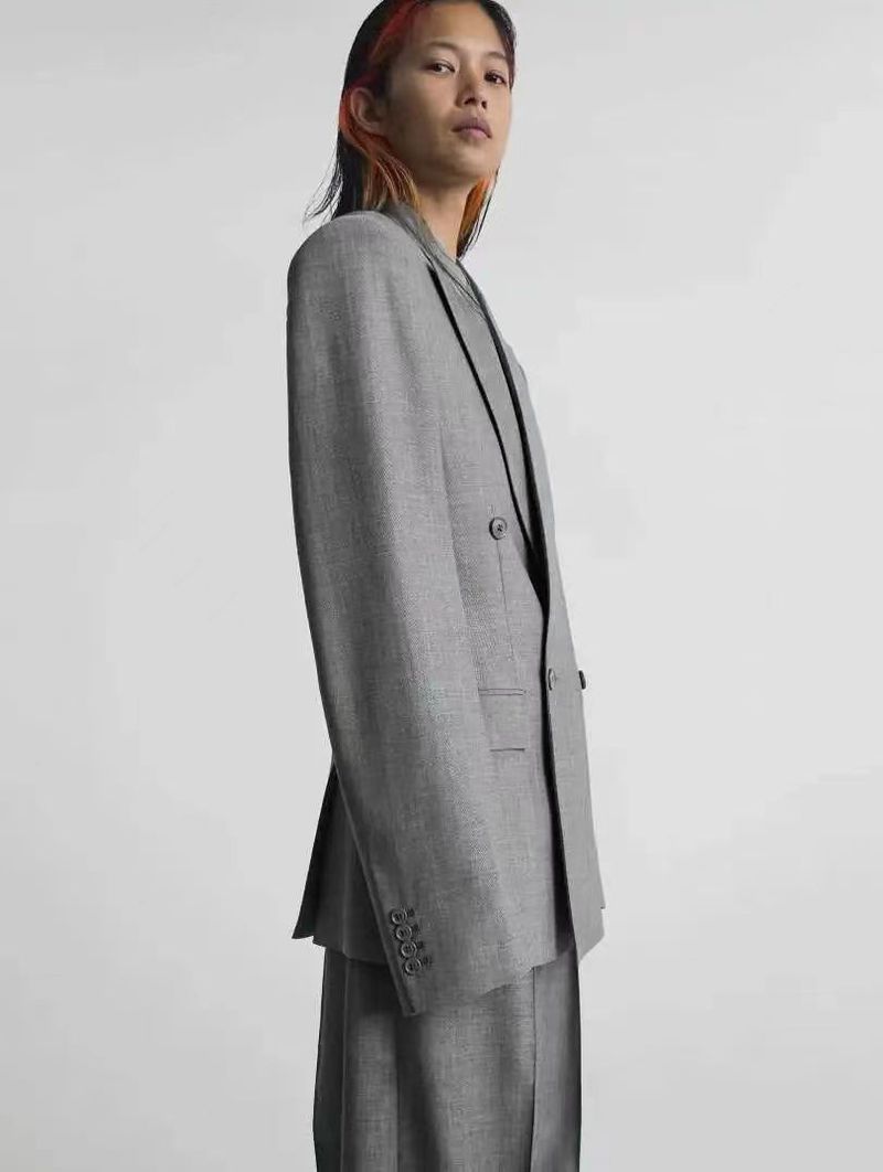 Model Karmay Ngai posing in profile wearing a tailored grey double-breasted blazer and matching trousers against a pale background.