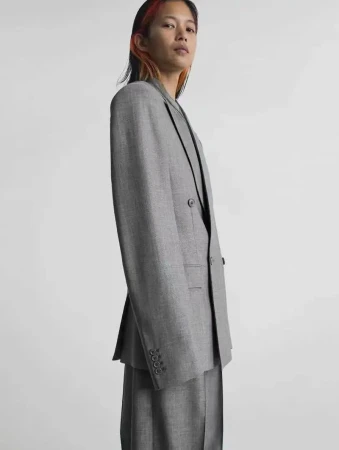 Model Karmay Ngai posing in profile wearing a tailored grey double-breasted blazer and matching trousers against a pale background.