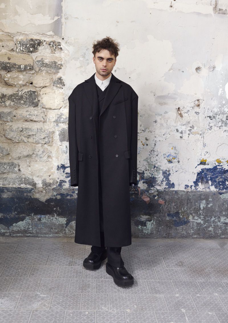 Young man in a stylish black coat stands confidently against a textured wall, showcasing a sophisticated fashion ensemble with platform shoes.