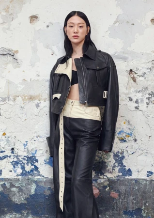Asian woman in a black leather jacket and black leather pants posing against a weathered wall in an eye-level medium shot.