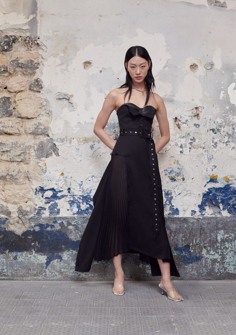 Elegant Asian woman poses in a black dress with a pleated skirt against a textured wall in a fashion editorial photograph.