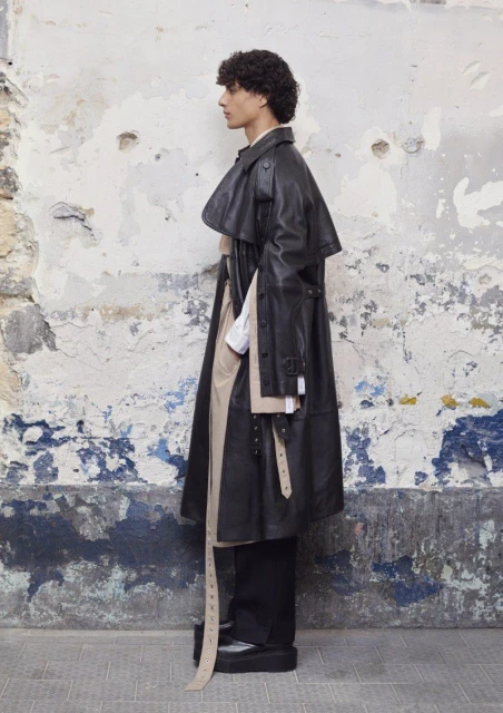 Male model wears a black leather trench coat in a side profile shot against a textured white and blue wall outdoors.