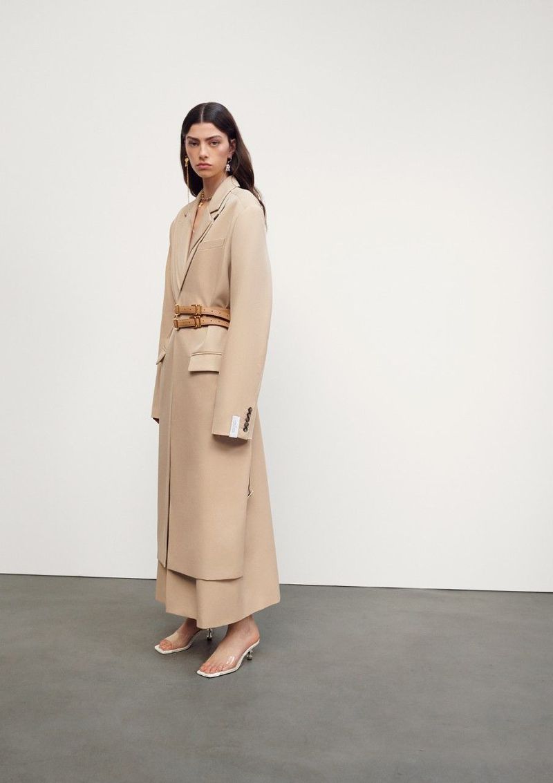 Elegant adult woman in beige trench coat and clear heels poses in a brightly lit studio against a neutral background for fashion editorial.