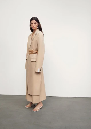 Elegant adult woman in beige trench coat and clear heels poses in a brightly lit studio against a neutral background for fashion editorial.