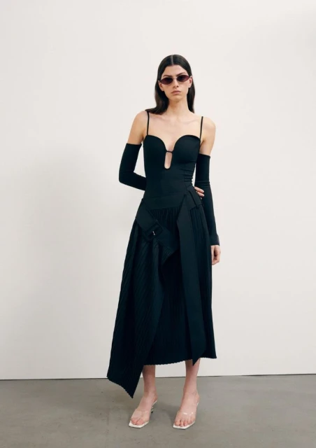 Elegant adult female fashion model posing in a studio wearing a stylish black dress and clear high heels against a white background.