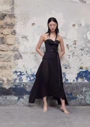 Model wearing a black strapless belted dress standing against a weathered stone wall for H&M x Rokh 2024 collection.