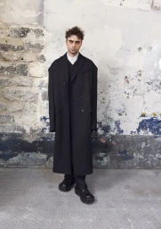 Model Lennon Gallagher poses wearing an oversized black double-breasted long coat and chunky platform shoes against a distressed wall.