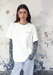 Model wearing a white t-shirt, blue floral sleeves, and light wash jeans standing against a distressed plaster wall