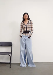 Model wearing a patterned cardigan and wide-leg jeans standing in a studio with a dark chair.
