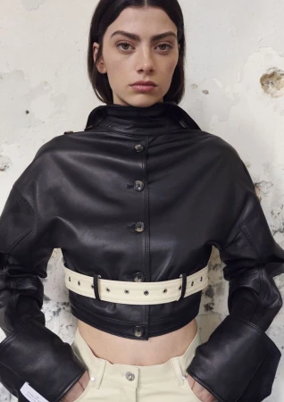 Model wearing a cropped black leather jacket with a cream eyelet belt and pale pants against a textured plaster wall.