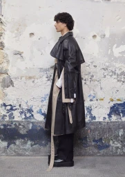 Full-length side profile of a male model wearing an oversized black leather trench coat against a distressed peeling wall.