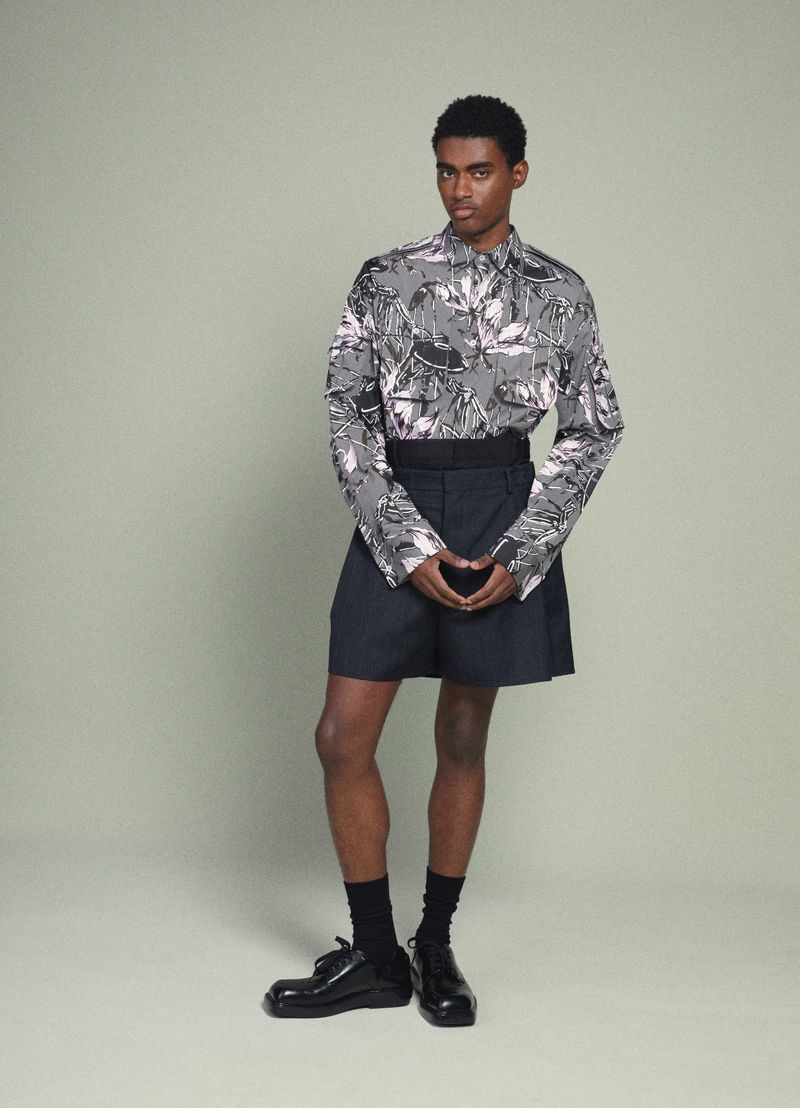 Man in patterned shirt and shorts stands in studio with clasped hands, wearing socks and dress shoes, looking at the camera.