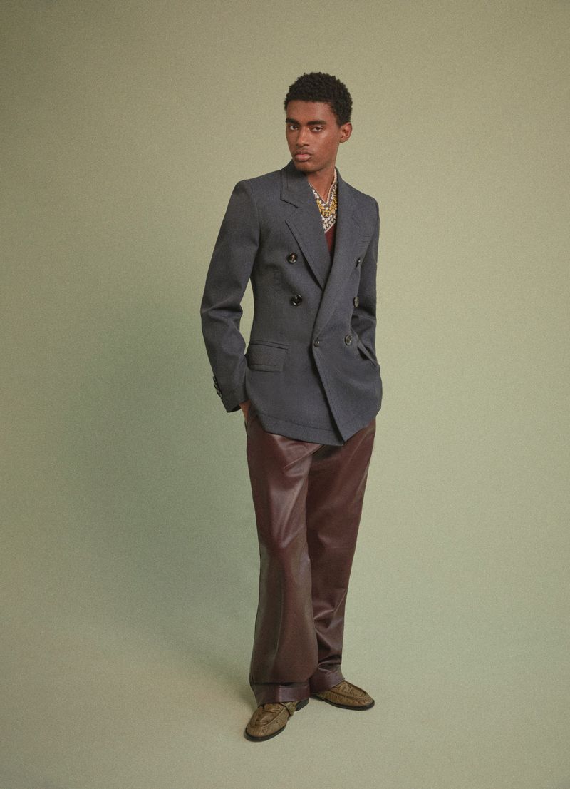 Full-length shot of a young adult male model posing indoors, wearing a gray double-breasted blazer, brown leather pants, and patterned shirt.