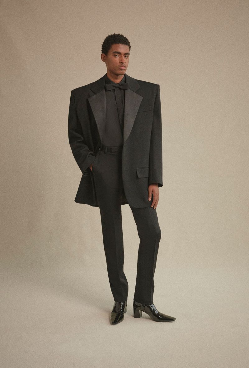 Man in oversized black tuxedo suit with bow tie and patent leather heeled shoes poses against a light tan background in a fashion studio.