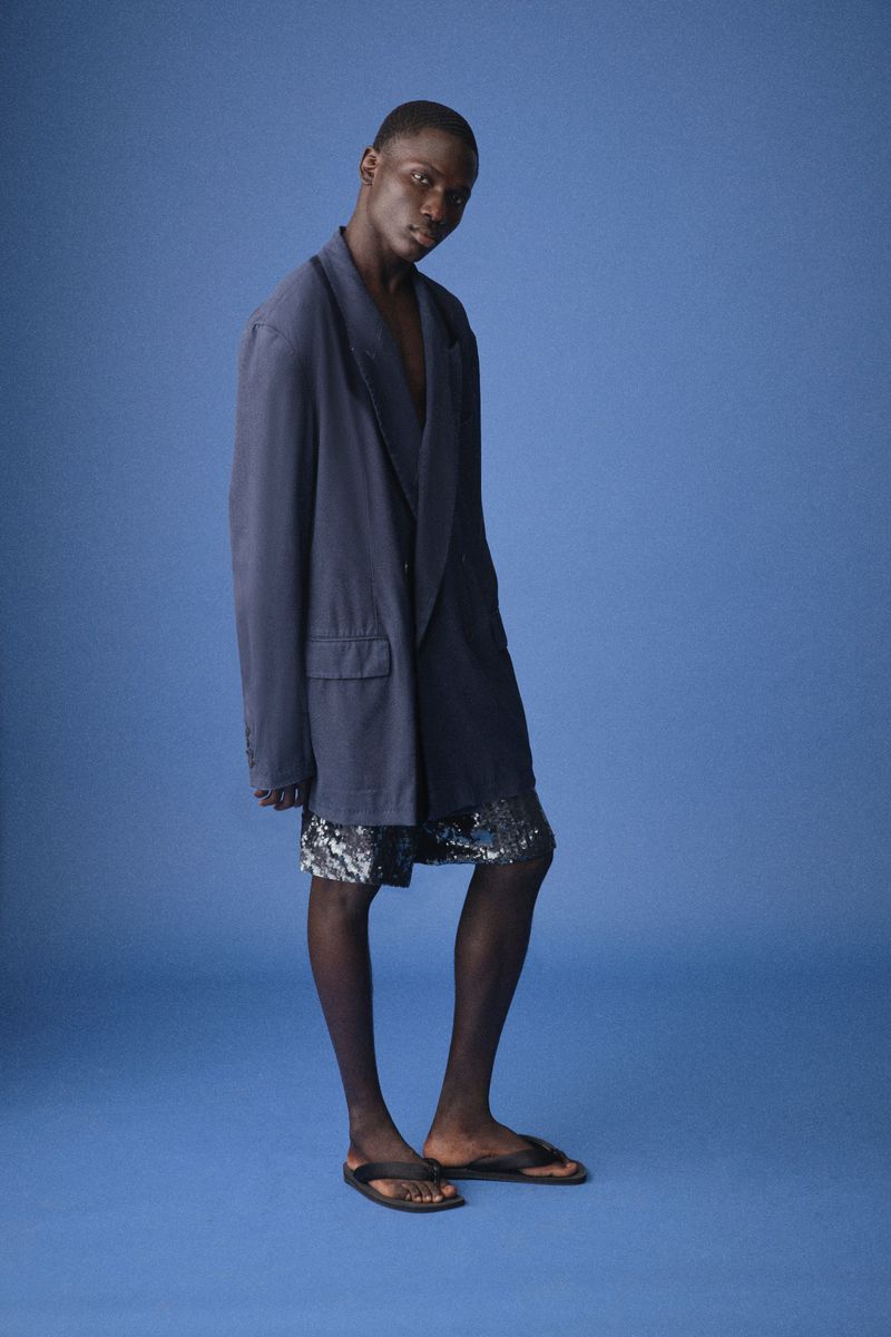 Dark-skinned man in blue blazer and sequined shorts poses in studio, wearing sandals against a bright blue backdrop.