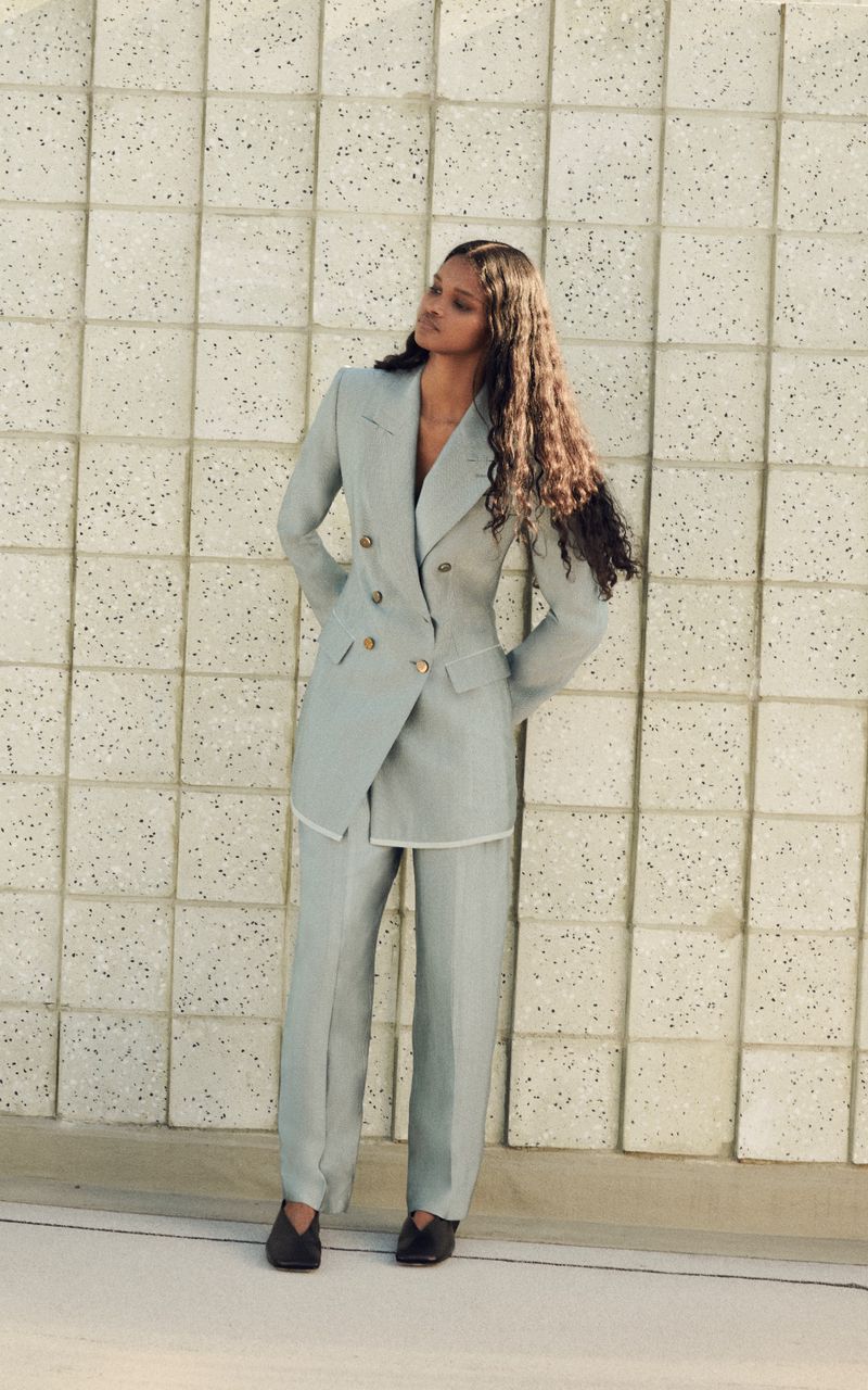 A woman poses in an elegant sage green suit against a textured wall, styled with long curly hair and dark shoes in a fashion shot.