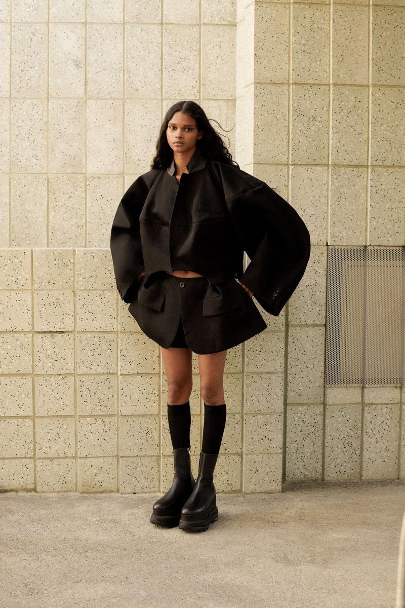 Fashionable woman in oversized black jacket and shorts standing against a tiled wall with chunky platform boots and knee socks.