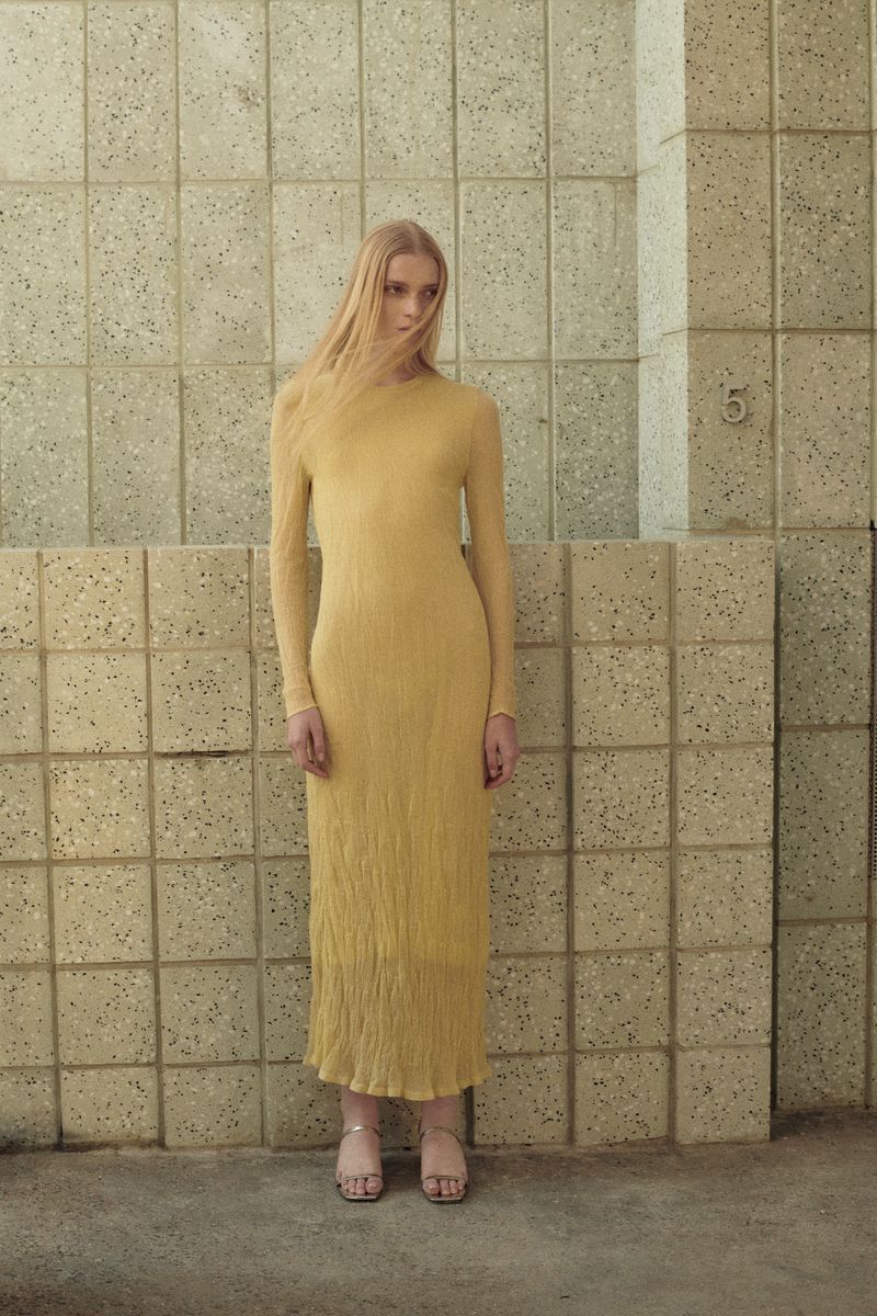 Full-length shot of a slender woman in a golden dress posing against a textured wall, showcasing fashion and neutral colors.