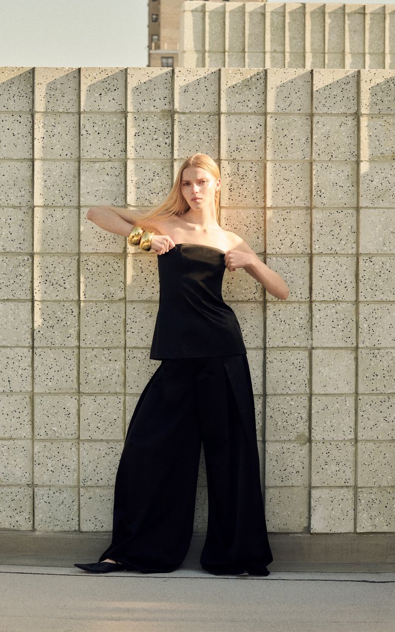A blonde woman poses in front of a textured wall wearing a black strapless top and wide-legged pants in a fashion shot.