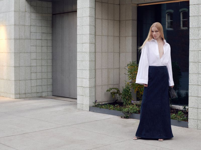 A fair-skinned woman with long blonde hair stands outdoors in an unbuttoned white shirt and navy maxi skirt against a building backdrop.