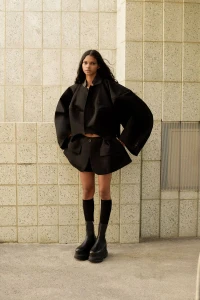 Model standing against a tiled stone pillar wearing an oversized black jacket and chunky boots