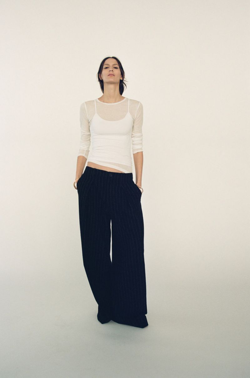 Full shot of a slender adult woman in a white top and dark, wide-leg pants against a bright white backdrop, looking directly at the camera.